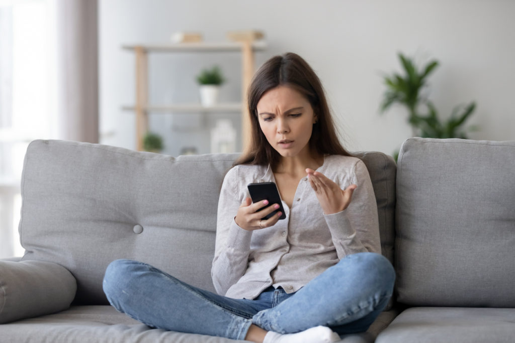 Indignant young woman sitting on couch in living room holding mobile phone
